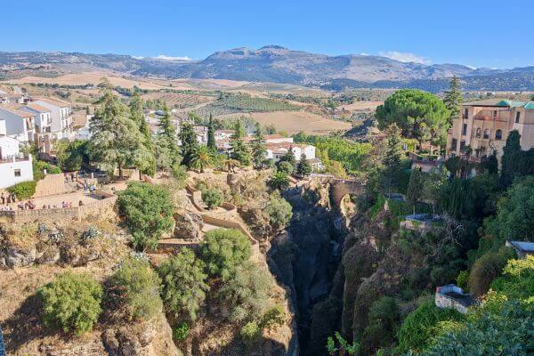 A view from Puente Nuevo Bridge, Ronda View from Puente Nuevo Bridge in Ronda showing the deep gorge with trees and vegetation, white buildings on both sides, and mountains in the distance under a blue sunny sky.