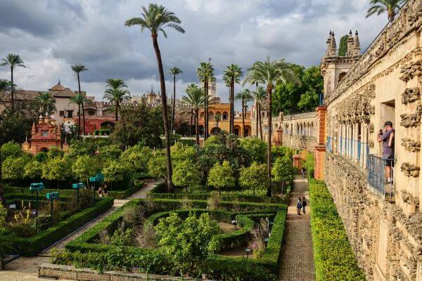 An elevated view of the gardens at the Royal Alcázar in Seville shows neat pathways, tall palm trees, and bright green hedges. Visitors walk through the garden while others look out from the stone balcony on the right. The scene highlights the peaceful layout of the historic palace grounds.