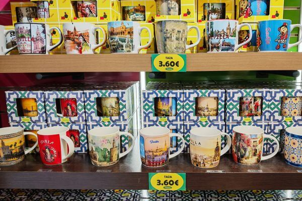 Souvenir mugs displayed on shelves in a Seville shop featuring colorful designs of Spanish landmarks and scenes, priced at 3.60 euros, with decorative tile backdrop.
