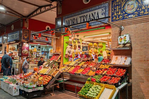 Mercado de Triana, Seville A colorful fruit and vegetable stall at Mercado de Triana in Seville, Spain. The stand is filled with fresh produce like tomatoes, bananas, peppers, and oranges, with a few shoppers browsing nearby. The bright sign above reads “Mora Triana No 45.”