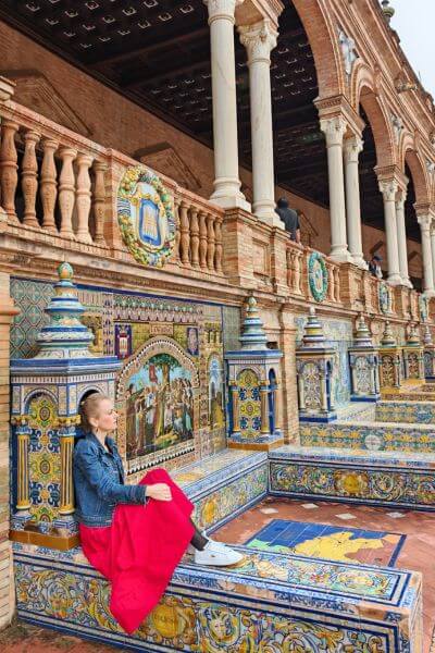 Plaza de Espana, Seville, Spain A woman sits on a colorful tiled bench at Plaza de España in Seville, Spain. The bench is decorated with detailed ceramic artwork and bright patterns beneath a row of arches and columns. She looks relaxed, enjoying the view of the beautiful plaza.