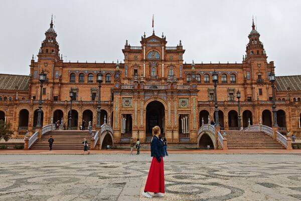 A woman in red skirt stands in the open plaza facing the grand brick building of Plaza de España in Seville. The wide courtyard leads up to the building’s arches, stairways, and towers, with a few visitors walking around. The scene highlights the scale and beauty of the landmark.
