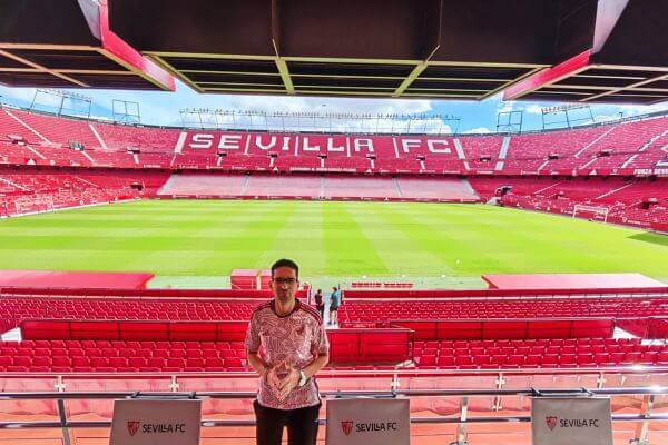 Man standing at Ramón Sánchez Pizjuán Stadium in Seville with red seats and green pitch visible behind him