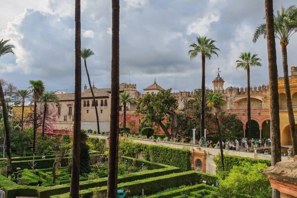 Royal Alcazar, Seville, Spain View of the Royal Alcazar gardens in Seville with tall palm trees, green hedges, and historic palace buildings under a cloudy sky