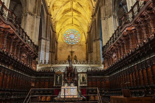 The photo shows the interior of Seville Cathedral with rows of dark wooden choir stalls lining both sides. A bright rose window glows above the altar area, filling the tall stone arches with warm light. The scene highlights the quiet and grand feel of the cathedral’s center space.