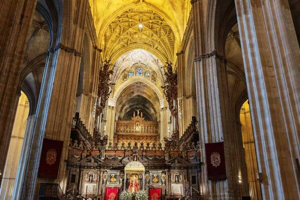 Seville Cathedral The main altar inside Seville Cathedral in Spain, with tall stone columns and a high arched ceiling glowing with warm light. A statue of the Virgin Mary stands at the center, surrounded by red banners and intricate woodwork. The space feels grand and peaceful.