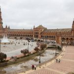 Wide view of Plaza de España in Seville with its semicircular brick buildings, central fountain, canal with bridges, and visitors walking across the tiled plaza under a cloudy sky