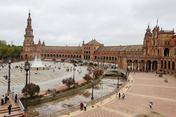 Wide view of Plaza de España in Seville with its semicircular brick buildings, central fountain, canal with bridges, and visitors walking across the tiled plaza under a cloudy sky