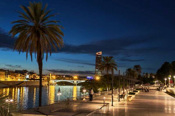 Evening walk along the Guadalquivir river in Seville with palm trees, lit streetlights, people strolling on the waterfront promenade, and Triana bridge in the background under a blue twilight sky.