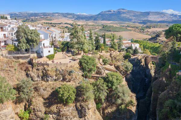 View from Puente Nuevo Bridge in Ronda showing the deep gorge with trees, Puente Viejo bridge below, white buildings on the cliff edge, and rolling hills with mountains in the distance.