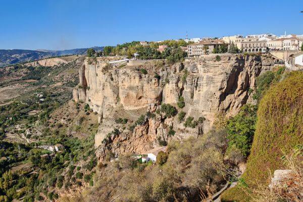 View of the deep gorge in Ronda, Spain with steep cliffs, white buildings on top, and part of Puente Nuevo Bridge visible on the right side.