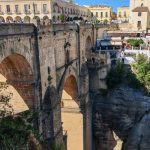 A wide stone bridge called Puente Nuevo spans a deep rocky gorge in Ronda, Spain, with sunlight highlighting its arches. People walk along the top of the bridge, and nearby buildings and outdoor cafes sit close to the cliff’s edge. The scene shows how the bridge connects both sides of the town over the dramatic drop.