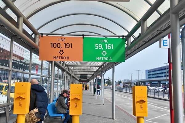 A bus stop at Prague Airport showing two signs - an orange sign for bus lines 100 and 191 connecting to Metro B, and a green sign for bus lines 59 and 161 connecting to Metro A, with passengers waiting on the benches and yellow ticket validators on the platform.