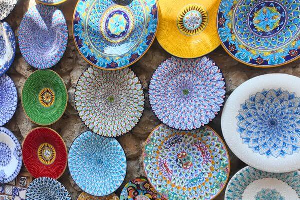 A display of colorful hand painted ceramic plates for sale on a stone wall on the Amalfi Coast, featuring blue, green, red and yellow patterns and flower designs.