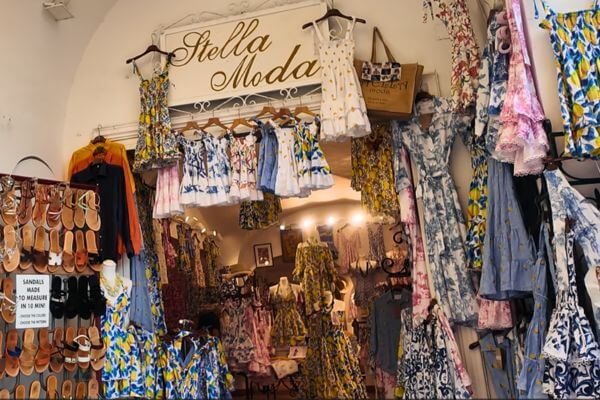 A colorful clothing shop called Stella Moda on the Amalfi Coast, with summer dresses in lemon and floral prints hanging on display, and handmade sandals on the wall with a sign saying sandals are made to measure in 15 minutes.