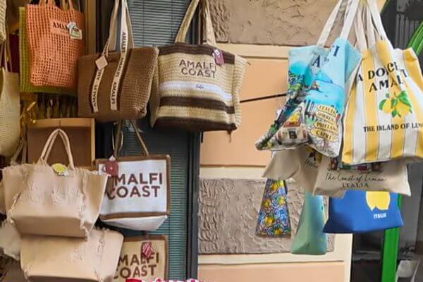 A display of Amalfi Coast souvenir handbags and tote bags for sale outside a shop, with lemon prints and Amalfi Coast writing on them.
