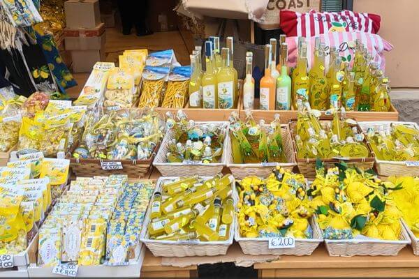 A market stall on the Amalfi Coast packed with lemon themed products including limoncello bottles, lemon candies, soaps and other lemon souvenirs displayed in baskets and on shelves.