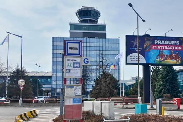 The Terminal 1 stop for the Airport Express AE bus at Václav Havel Airport Prague, with a bus stop sign, the airport control tower in the background, and a billboard advertising Aquapalace on the right.