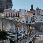 Cars parked along the waterfront in Atrani town on the Amalfi Coast, Italy, with a road running above on stone arches and the church and hillside buildings in the background.