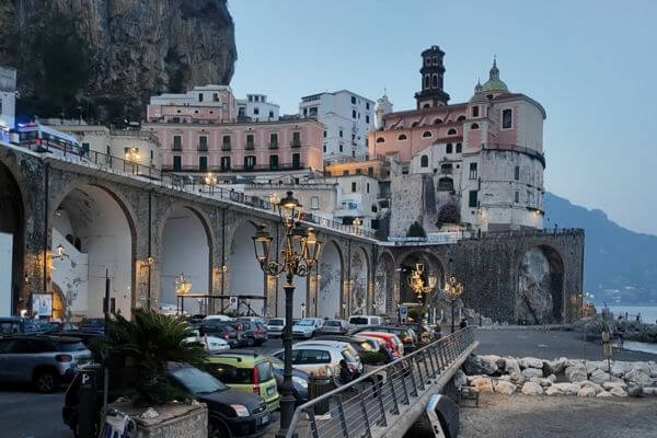 Cars parked along the waterfront in Atrani town on the Amalfi Coast, Italy, with a road running above on stone arches and the church and hillside buildings in the background.