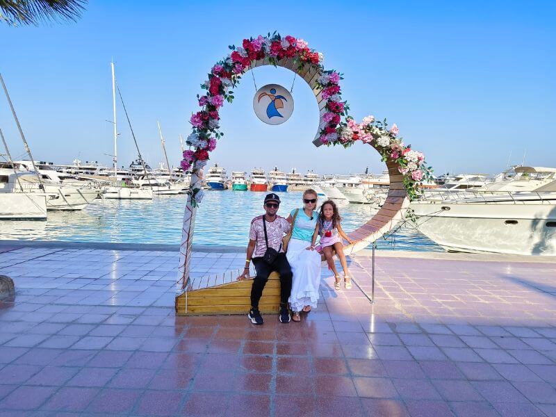 A family posing by a flower arch at Hurghada Marina in Egypt, with boats and yachts in the background.